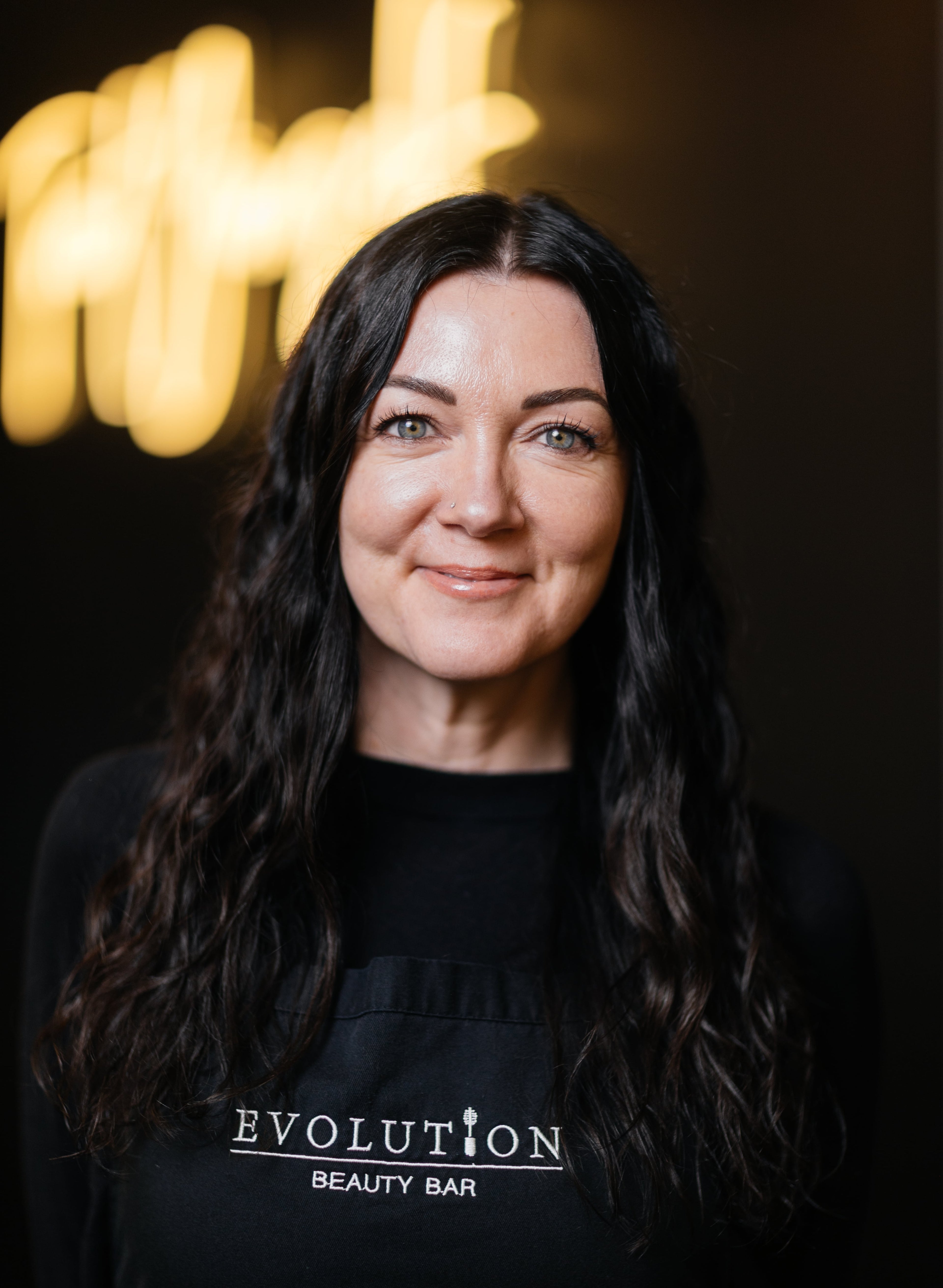 Woman wearing a black apron with 'Evolution Beauty Bar' logo against a dark background.